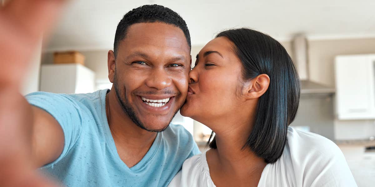 "She's my forever." A loving couple taking a selfie together at home. He smiles proudly, she kisses his check.