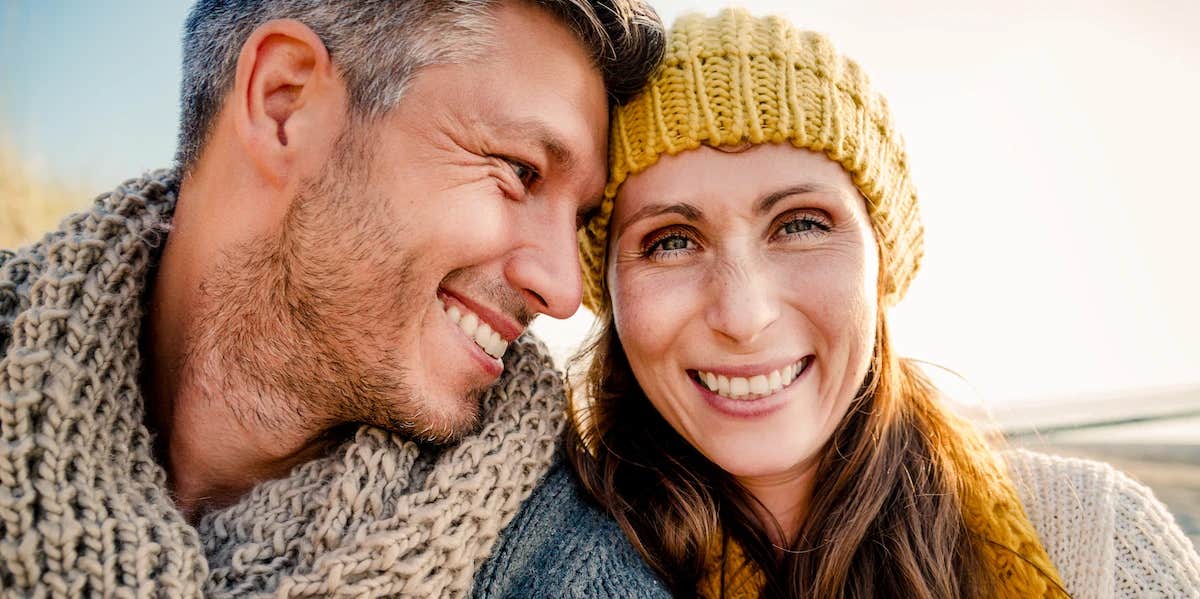 a woman looks at the camera smiling while a man lovingly rest his forehead on the side of her head=