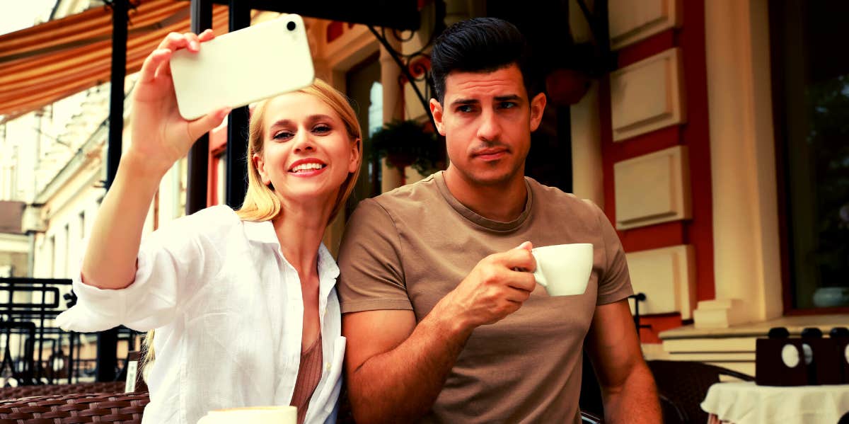happy woman taking selfie of herself and her unhappy boyfriend at a coffee shop 