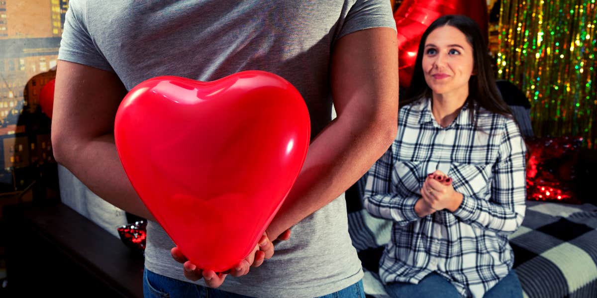 man approaching woman while holding big red plastic heart between his hands behind his back to present to her