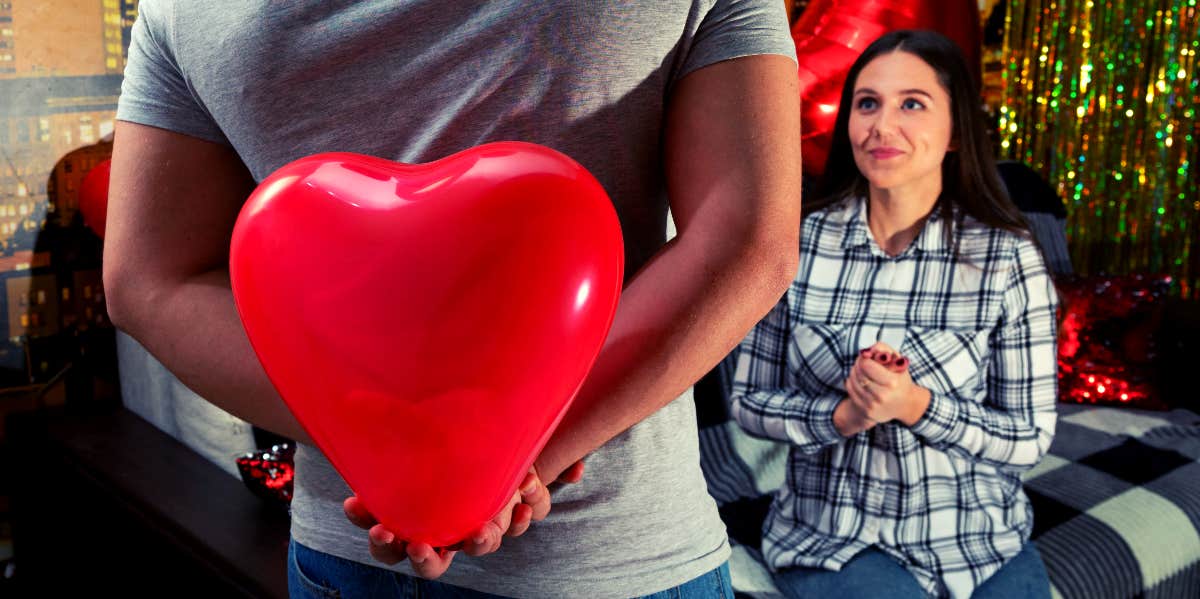 How To Know If Someone Is Interested In You: 14 Signs They're Into You man approaching woman while holding big red plastic heart between his hands behind his back to present to her