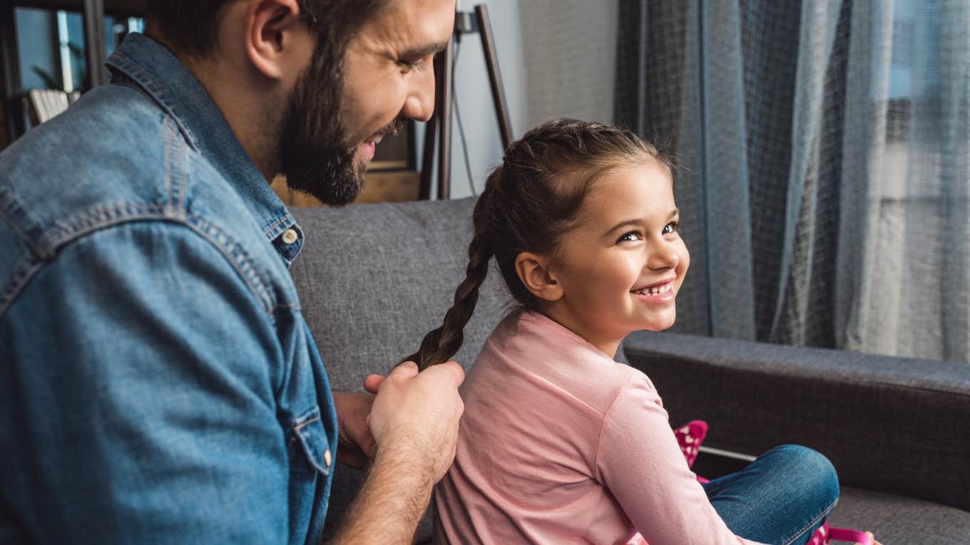 father braiding his daughter's hair while sitting on couch at home