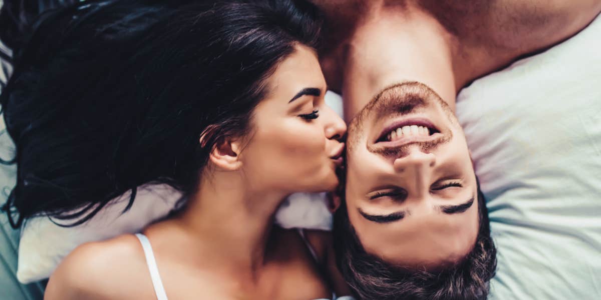 young adult couple with brown hair reclines on bed, him upside down, while she kisses his cheek