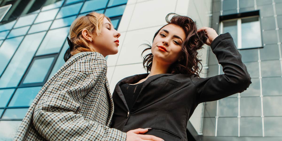 two young women embrace in front of a tall building, one is happy, one is not