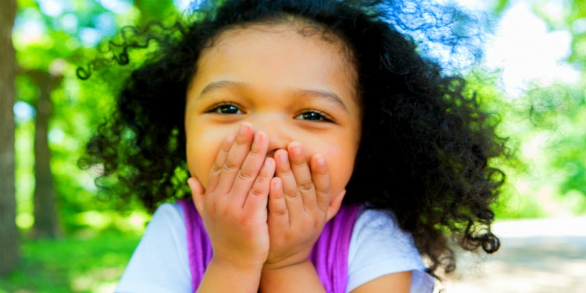 young girl with brown skin and black hair covers her mouth and grins