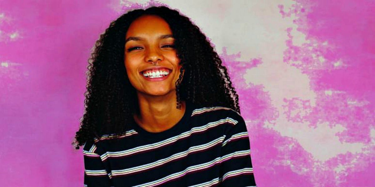 6 Ways To Stop Making Excuses When You Keep Giving Up On Your Dreams young woman with dark skin and black hair smiles broadly in front of pink background