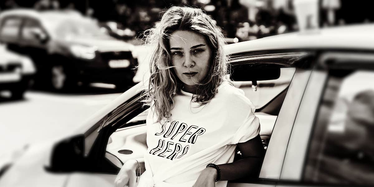 young woman with an angry expression leans back against a car in the city 