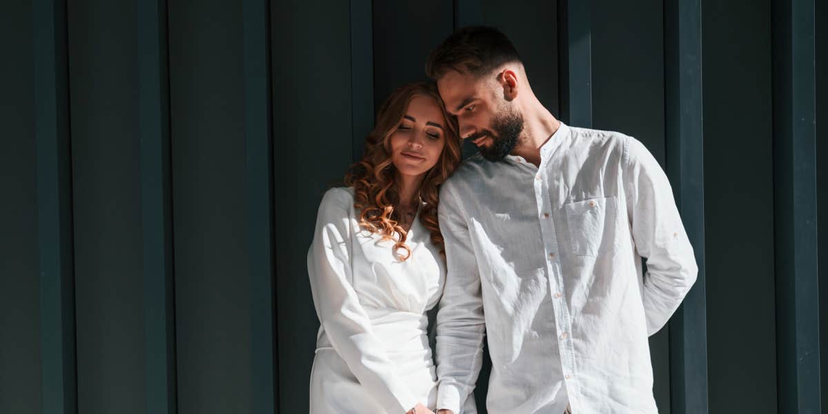 Couple in white clothing gently snuggling in front of a dark green wall