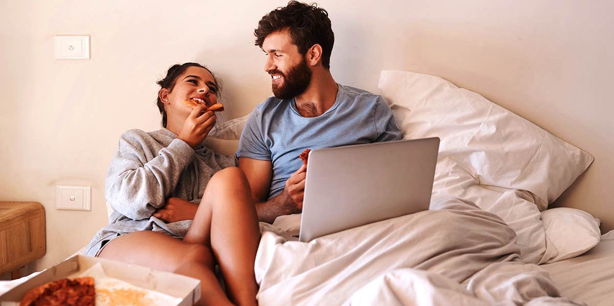 couple sitting in guy's bed with laptop and pizza