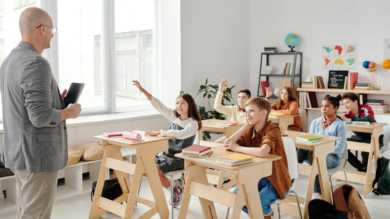 teacher standing in the front of classroom while students raise their hands at desks