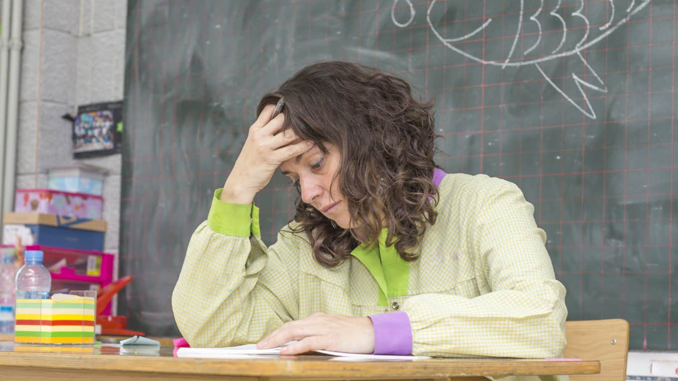 Teacher Questions Her Job After A Parent Of A Child Who Kicked & Punched Her During A Tantrum Threatened To Sue stressed teacher with head in hand sitting at desk in classroom