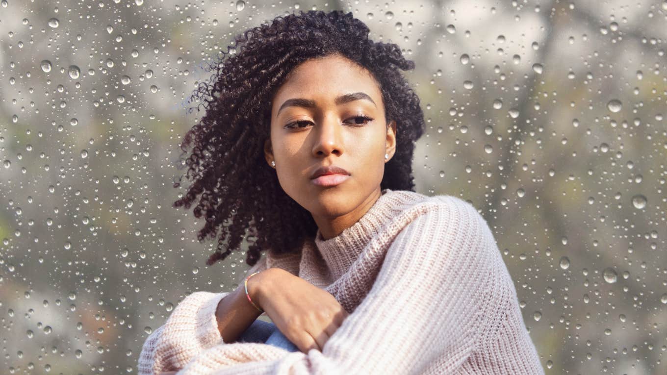 Woman looking solemn next to a rainy window. 