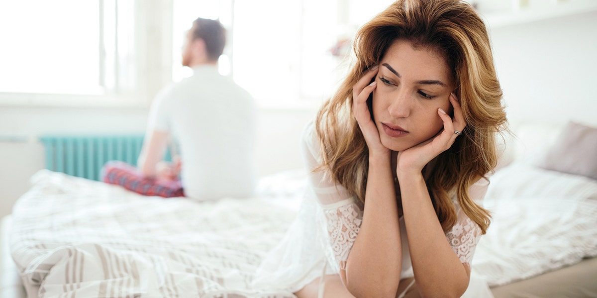woman looking forlorn on bed with husband in background