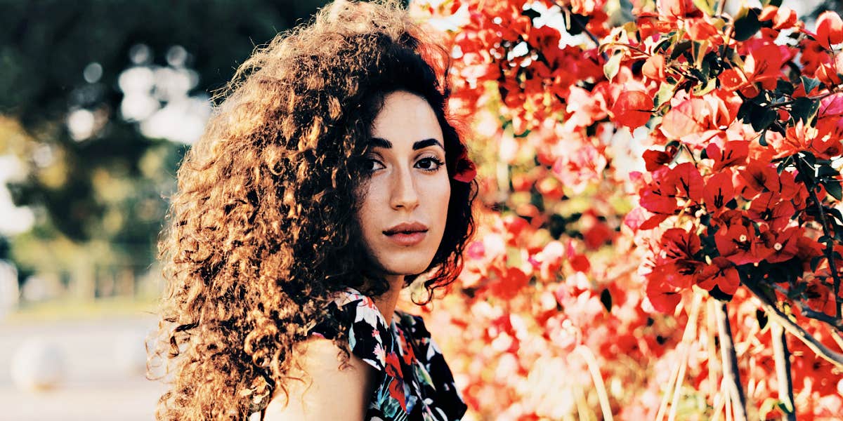  woman with curly dark hair standing in front of pink bushes