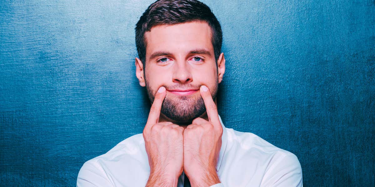 man in front of blue background trying to make himself smile with his fingers