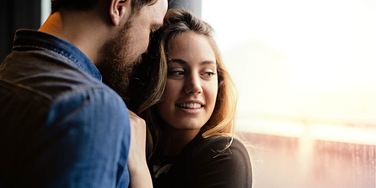 couple standing near window, him looking at her while she looks out the window, half-smiling