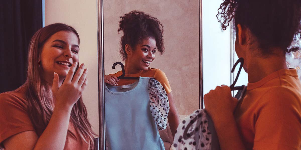 A photograph of two women looking into a mirror, with one holding up a blouse to herself with a grin.