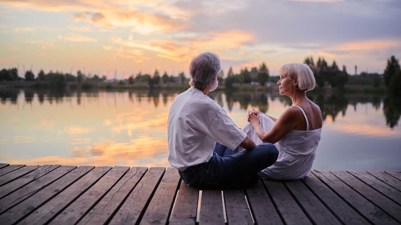 middle-aged couple sitting together on lake bank enjoying beautiful sunset