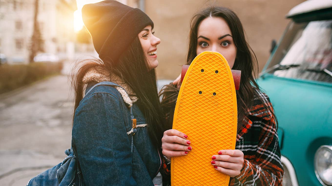 Two street girls smiling and joking with skateboards near vintage cars