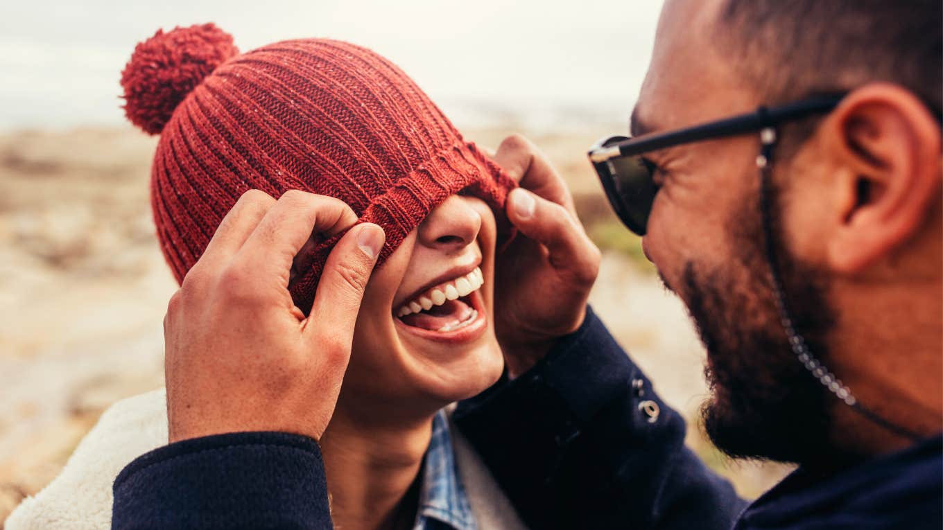 Loving couple having fun outdoors. Man covering eyes of woman with cap.