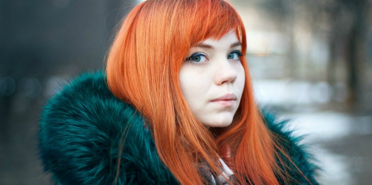 young woman with very pale skin and bright red hair looks sideways at the camera in a green faux fur jacket