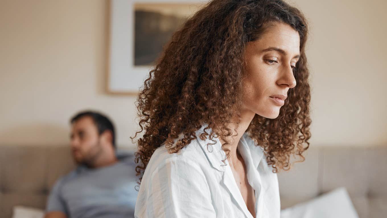 upset woman sitting at edge of bed after fight with husband lying behind her