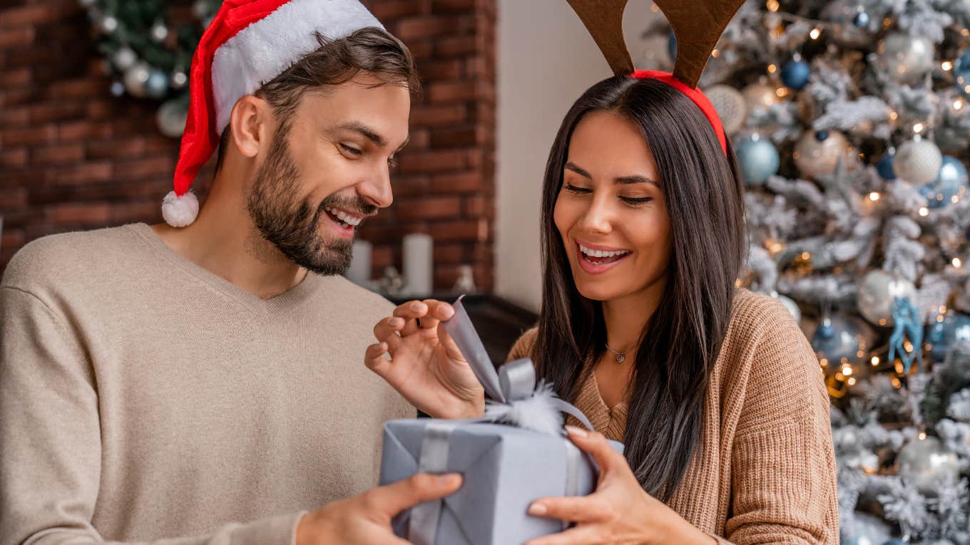 spouse giving Christmas gift to his girlfriend sitting beside a Christmas tree at home.