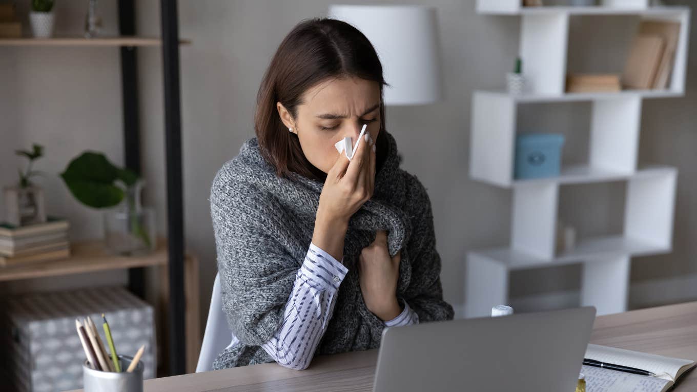woman blowing nose with tissue while sitting in front of her laptop