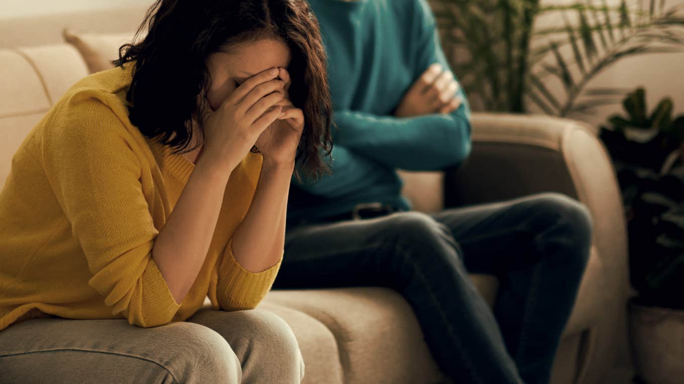 woman sitting on couch with face covered while husband sits with arms crossed