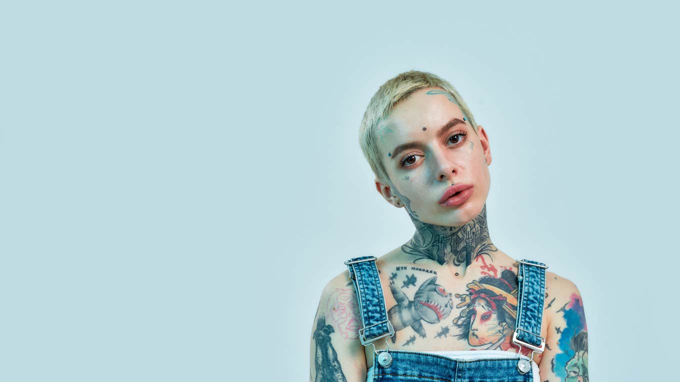  tattooed and pierced white girl wearing denim overall standing and looking into a camera on a light blue color background