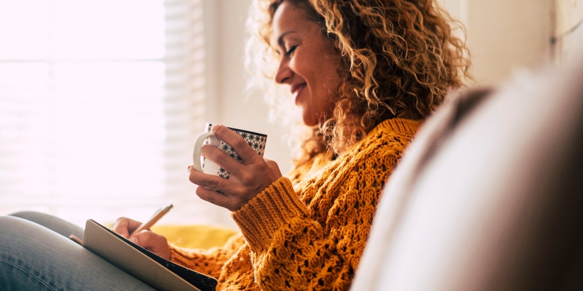 woman writing on the couch while holding a mug