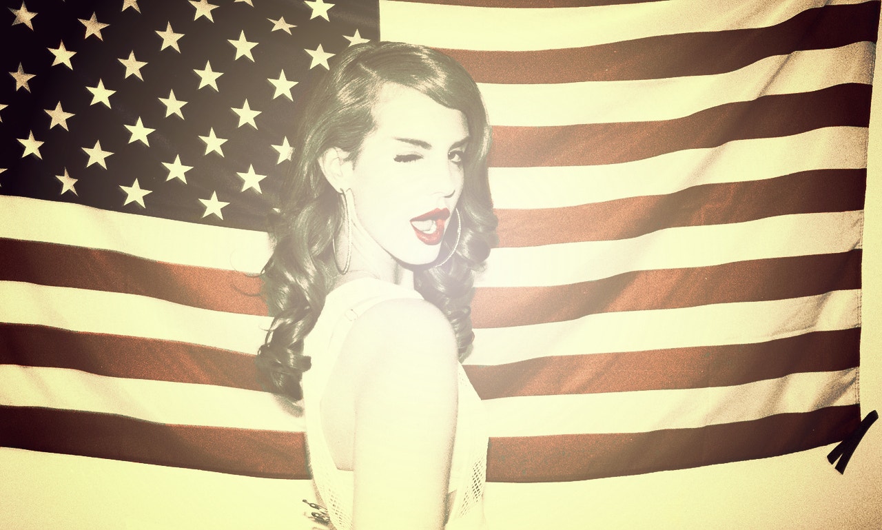 Lana Del Rey winking wearing red lipstick in front of the United States American flag for her "Ride" music video