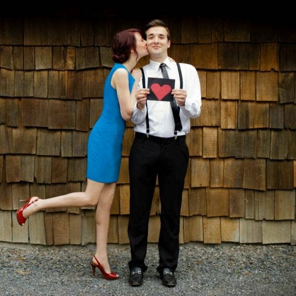 a hopeful romantic couple kiss, holding a heart shape on a card