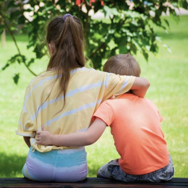 Older sister and younger brother sit on a bench at a park next to each other.