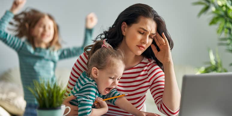 stressed mom working at desk with laptop while holding crying toddler with daughter jumping in background