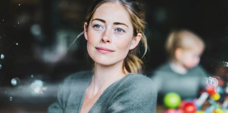 A calm woman gazes out the window while her child plays behind her, symbolizing self-care, emotional boundaries, and the power of learning to put herself first without guilt.