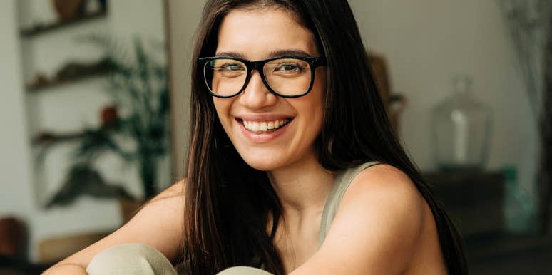 Smiling woman wearing glasses sitting comfortably indoors, appearing relaxed, confident, and emotionally grounded.
