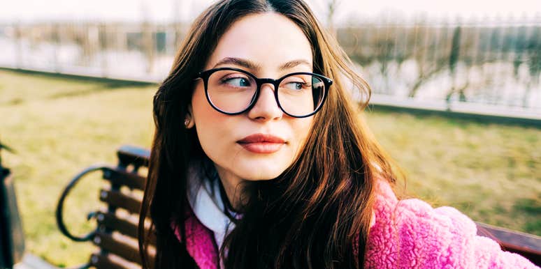 Woman wearing glasses sitting on a park bench and looking off to the side, appearing thoughtful and composed during a quiet moment of reflection.