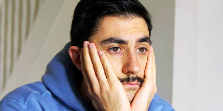 Man in a blue hoodie resting his face in his hands, staring ahead with a tired and emotionally drained expression.