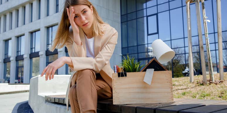 Frustrated woman sitting on bench with box of personal belongings near business center. 