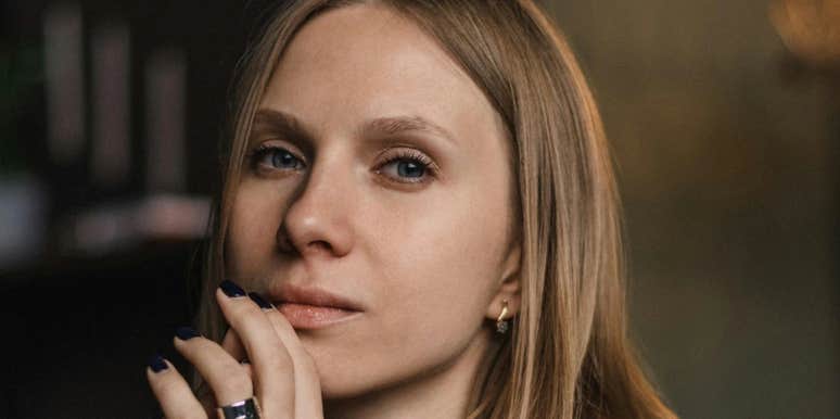 A woman rests her hand near her face while looking toward the camera indoors.