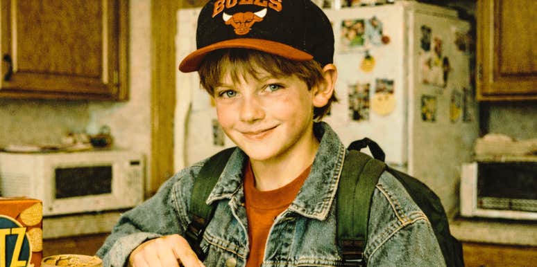 A young boy wearing a backpack and baseball cap eats a snack in a kitchen.