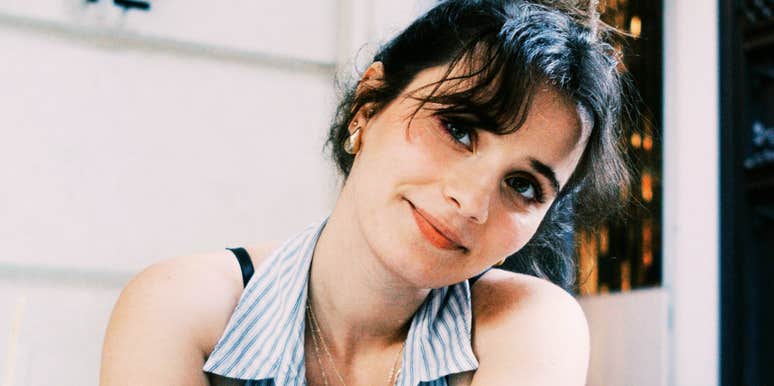 A close-up portrait of a woman with soft bangs and a gentle smile, seated indoors near a window.