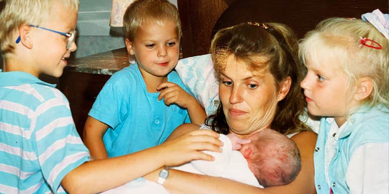 A mother holds a newborn while three young children gather closely around her indoors.