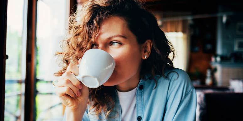 Woman with curly hair drinking coffee by a window in a cozy home setting