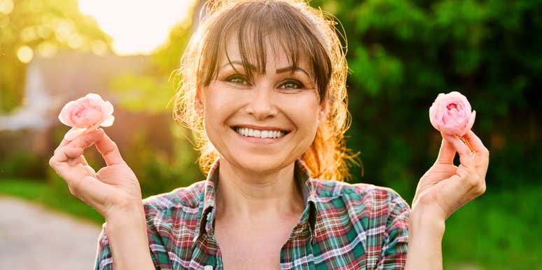 Smiling woman holding two pink flowers outdoors in natural light.