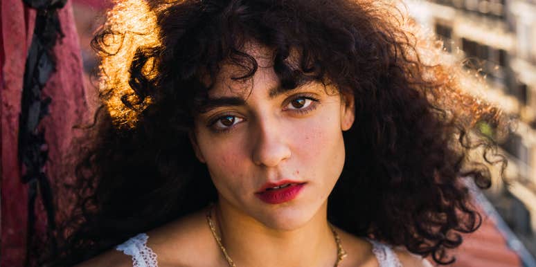 A close-up portrait of a woman with curly hair looking at the camera outdoors.