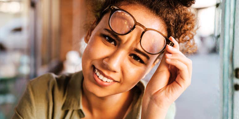 A woman smiling indoors wearing glasses, resting her chin on her hand.