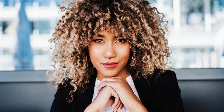 Woman with curly hair wearing a blazer, resting her chin on her hands and looking directly at the camera indoors.