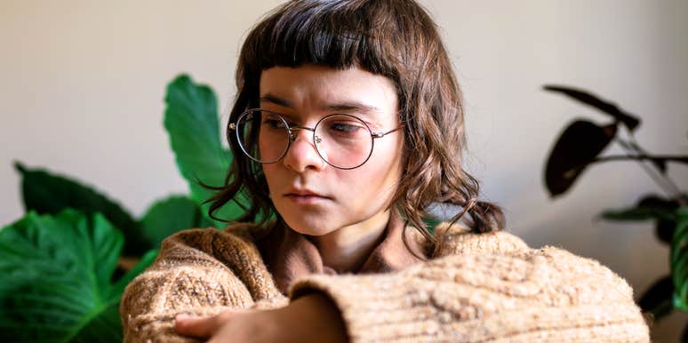 A person wearing glasses sits indoors with their arms folded, looking down thoughtfully.